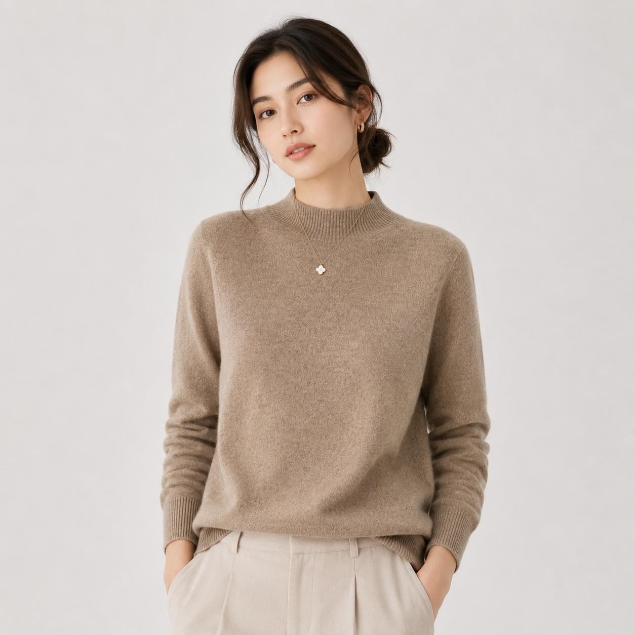 A woman wearing a light taupe knit mock-neck sweater and cream trousers, standing against a solid light gray background.