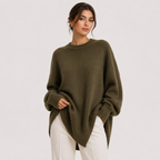 A woman wearing an oversized olive green knit sweater and white pants, standing against a plain light gray studio background.