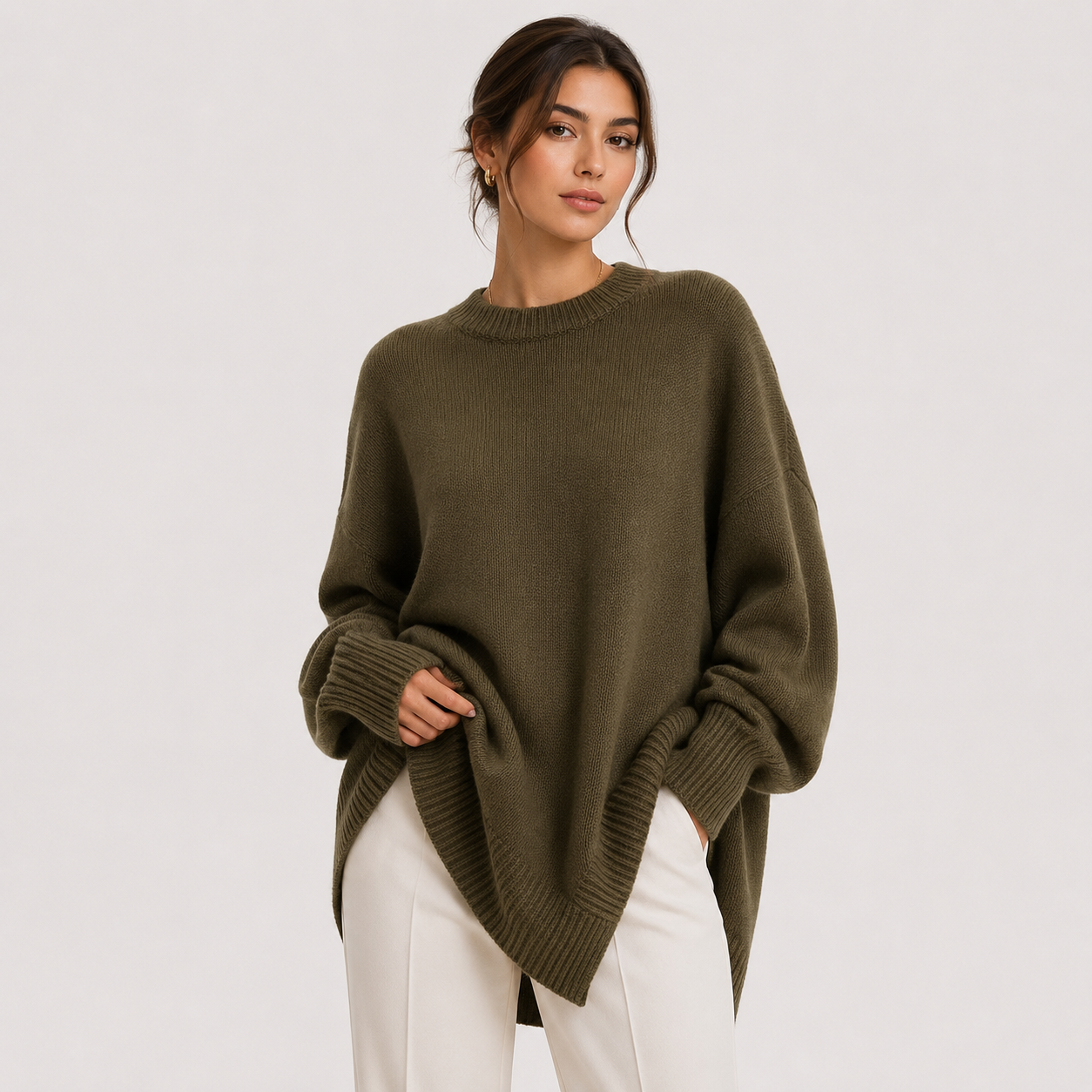 A woman wearing an oversized olive green knit sweater and white pants, standing against a plain light gray studio background.