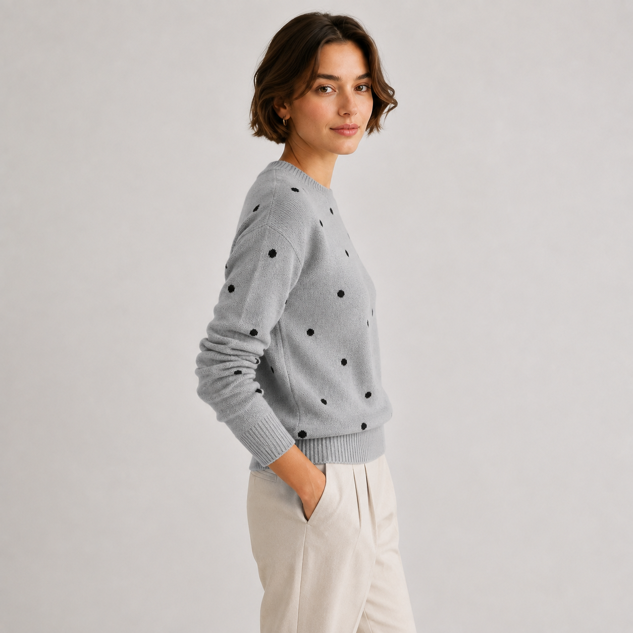 A woman wearing a light gray knit sweater with black polka dots and cream-colored trousers, standing against a light gray studio background.