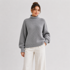 A woman wearing a gray chunky knit turtleneck sweater and white wide-leg trousers, standing against a light gray studio background.