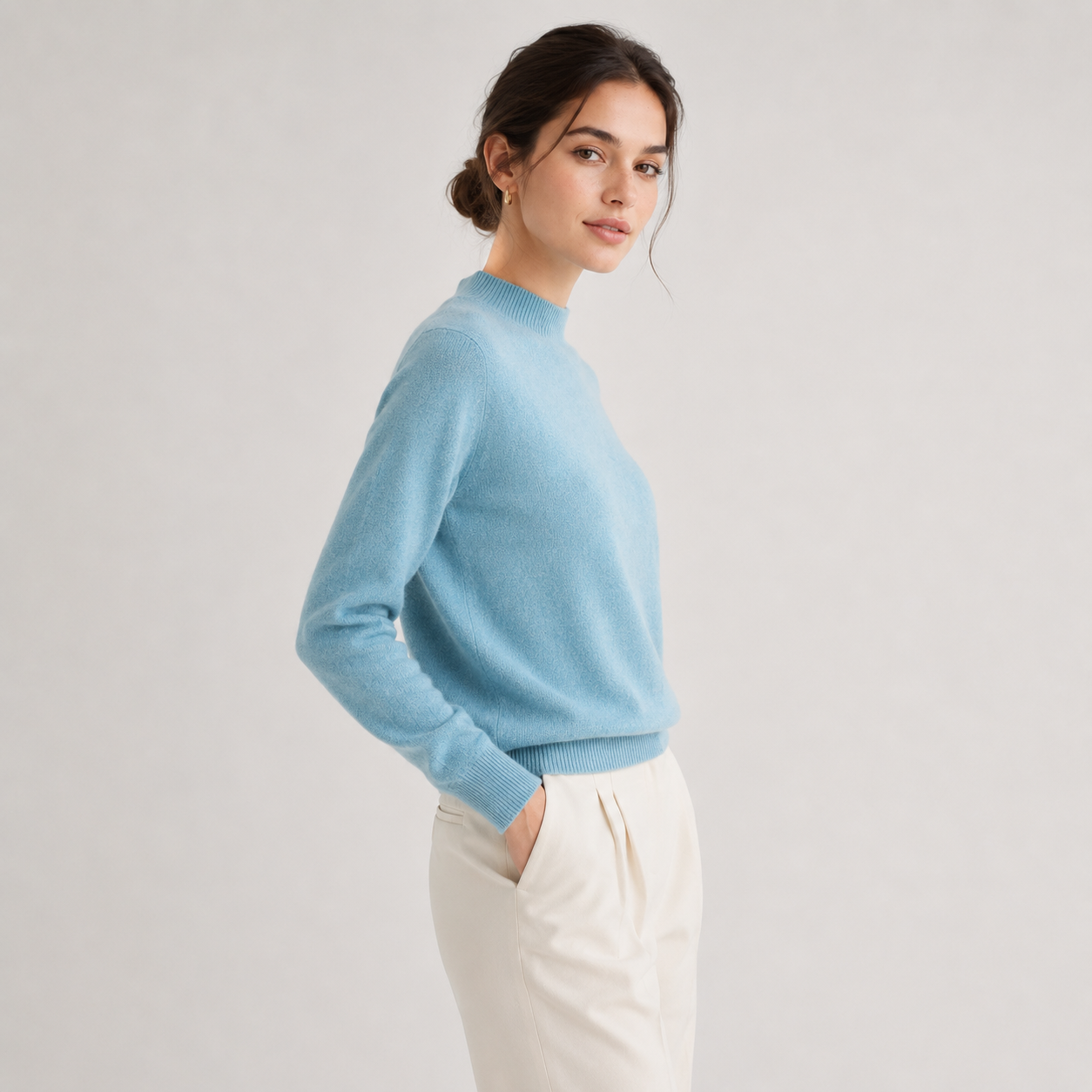 A woman wearing a light blue mock neck knit sweater and cream-colored trousers, standing against a neutral light gray studio background.