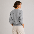 A woman shown from the back wearing a light gray knit sweater with black polka dots and cream pants, photographed against a plain light gray studio background.