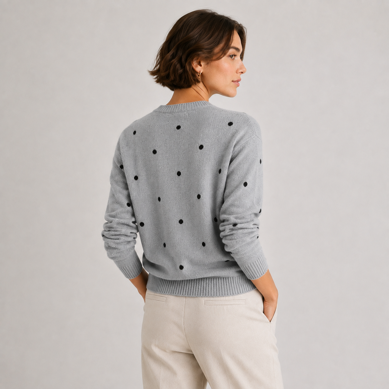 A woman shown from the back wearing a light gray knit sweater with black polka dots and cream pants, photographed against a plain light gray studio background.