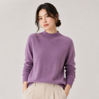 A woman wearing a soft lavender-purple knit mock neck sweater and light beige trousers, standing against a plain light gray studio background.