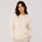 A woman wearing a cream-colored knit crewneck sweater and light beige trousers, standing against a plain light gray studio background.