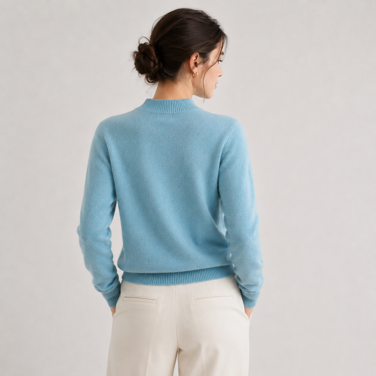 A woman shown from the back wearing a light blue knit sweater and cream trousers against a plain light gray studio background.
