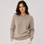 A woman wearing a light taupe knit crewneck sweater and white trousers, standing against a neutral light gray studio background.