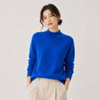 A woman wearing a bright royal blue mock-neck knit sweater and light beige trousers, standing against a plain light gray studio background.