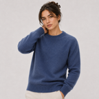 A woman wearing a blue crewneck knit sweater and cream pants, posing against a light gray studio background.