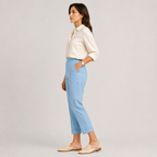 A woman stands in profile wearing a cream button-up shirt, light blue cropped trousers, and beige flat mules against a plain light gray studio background.