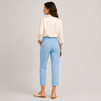 A woman is shown from the back wearing a cream-colored long-sleeve blouse and light blue cropped trousers, paired with beige flat sandals, against a neutral light gray studio background.