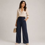 A woman stands against a light gray studio background wearing a cream satin button-up blouse, navy wide-leg drawstring pants, and beige heels, while holding a small beige handbag.