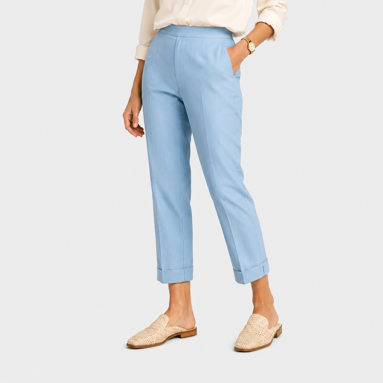A model wearing light blue cropped trousers with rolled cuffs, paired with a cream blouse and beige woven mule flats, shown against a light gray studio background.
