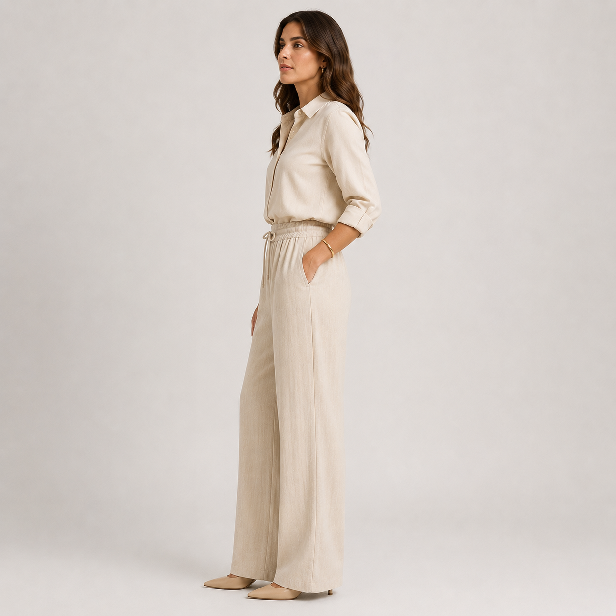 A woman stands in profile wearing a light beige linen-blend jumpsuit with a button-up shirt collar, long sleeves, and wide legs, paired with beige pointed flats, against a light gray studio background.