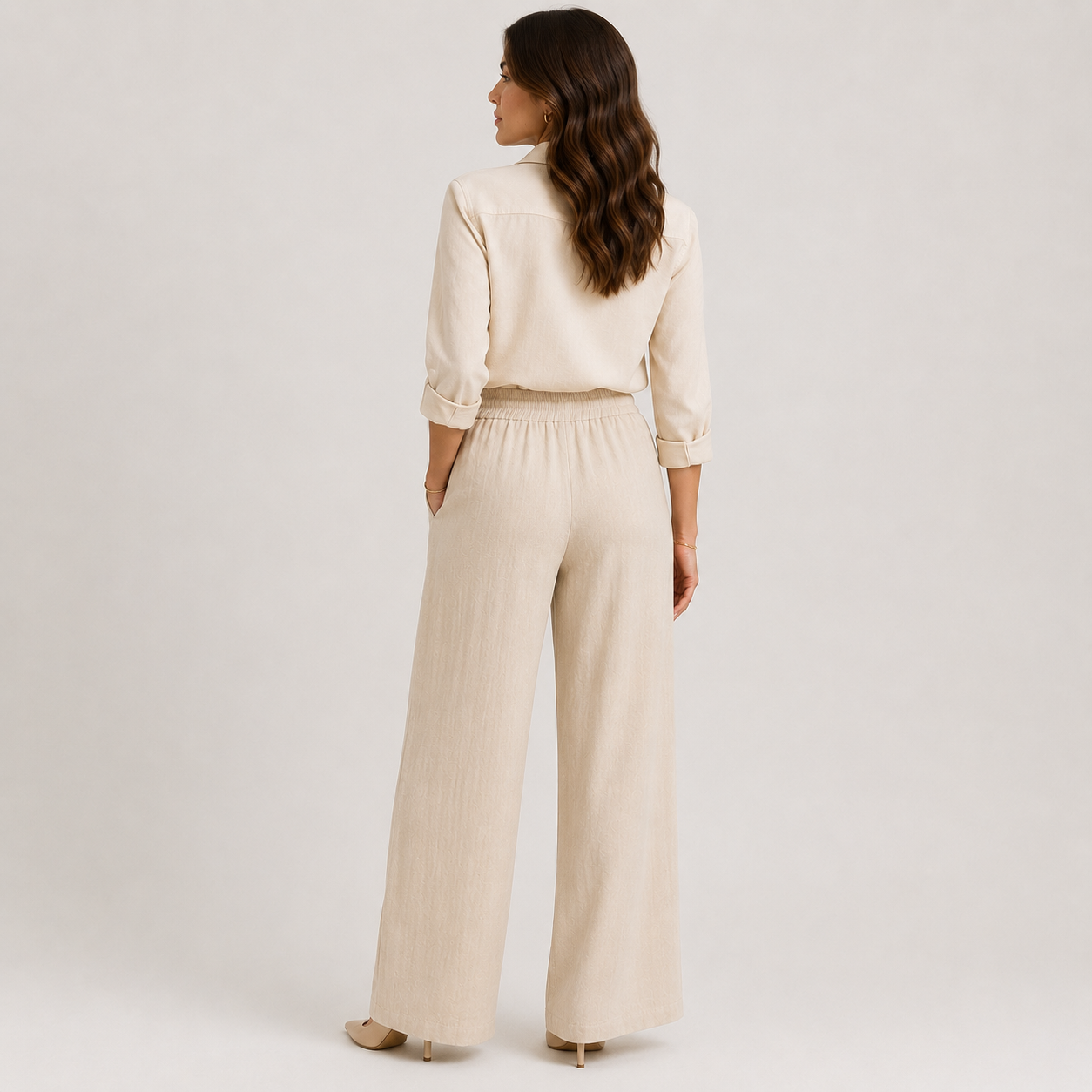A woman shown from the back wearing a matching cream-colored two-piece set consisting of a long-sleeve collared top and high-waisted wide-leg trousers, photographed against a neutral light gray studio background.