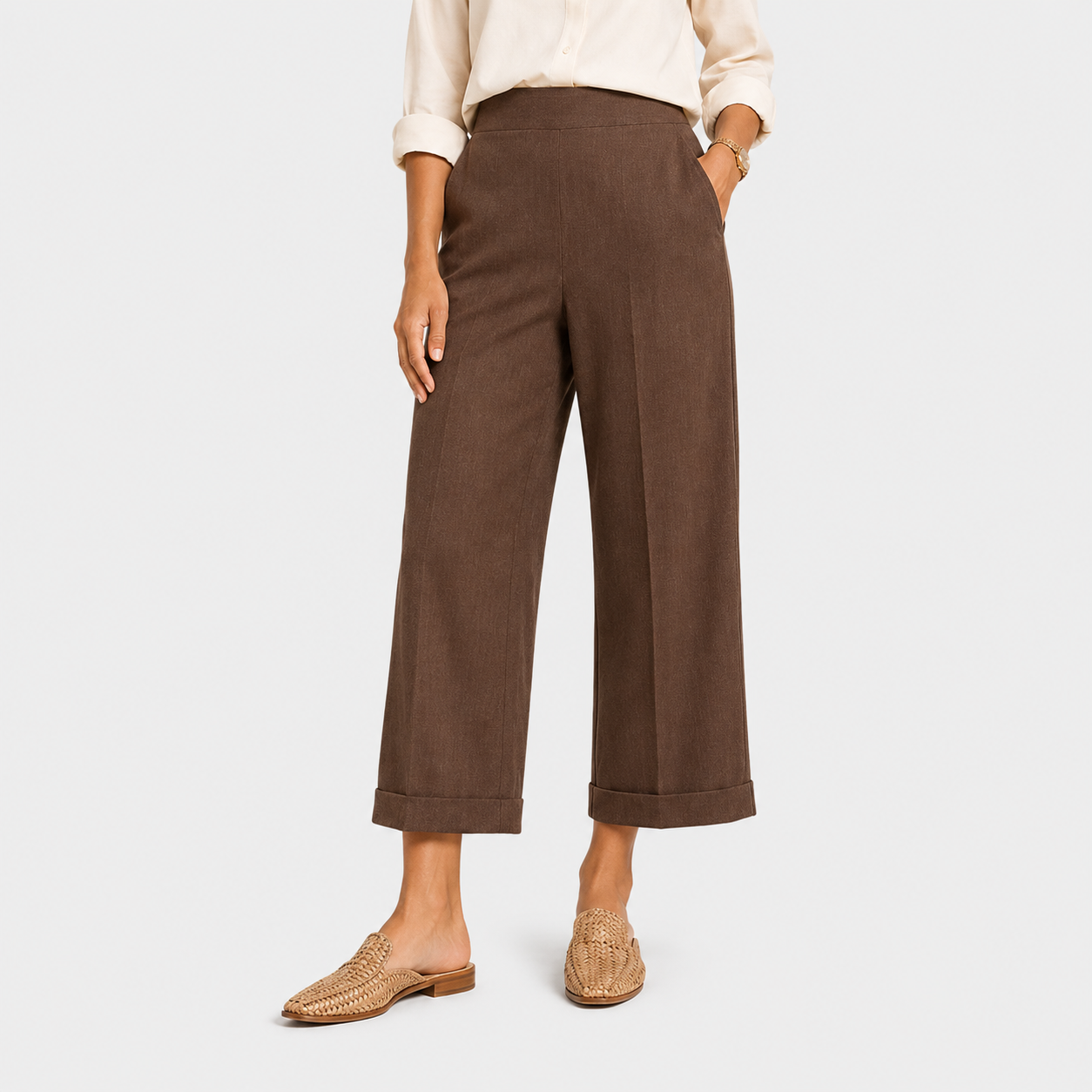 A model wearing brown wide-leg cropped trousers with a cream blouse and woven tan loafers, shown from the waist down against a light gray studio background.