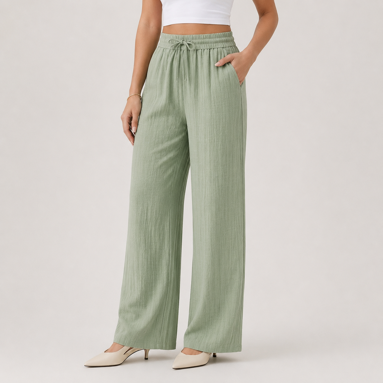 A model wearing sage green wide-leg pants with a drawstring waist, paired with a white crop top and beige pointed-toe heels, photographed against a neutral light gray studio background.