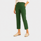 A model wearing a pair of cropped, straight-leg trousers in forest green, paired with a cream blouse and beige woven mule flats, set against a solid light gray background.