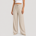 A model wearing high-waisted, wide-leg linen pants in a light beige color, paired with a white crop top and beige pointed-toe heels, set against a plain light gray studio background.
