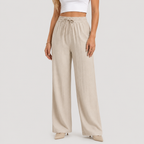 A model wearing high-waisted, wide-leg linen pants in a light beige color, paired with a white crop top and beige pointed-toe heels, set against a plain light gray studio background.
