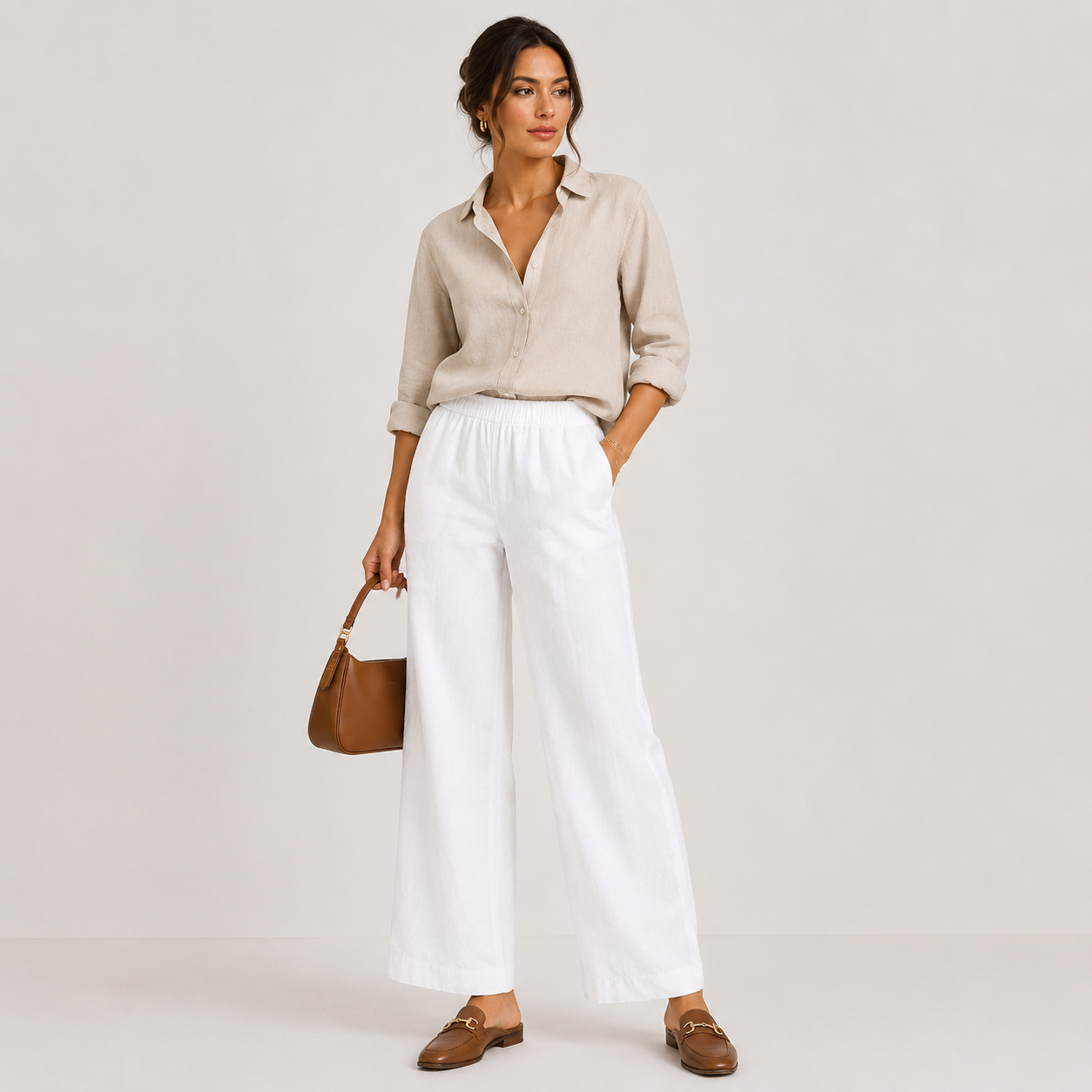 A woman stands against a light neutral studio background wearing a light beige linen button-up shirt, white wide-leg cropped pants, and brown loafers, while holding a brown handbag.