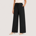 A model wearing black wide-leg pleated trousers with a drawstring waist, paired with a cream top and beige pointed-toe heels, against a light gray studio background.