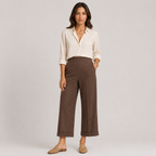 A woman stands against a light gray studio background wearing a cream button-up shirt, brown wide-leg cropped pants, and tan flat sandals.