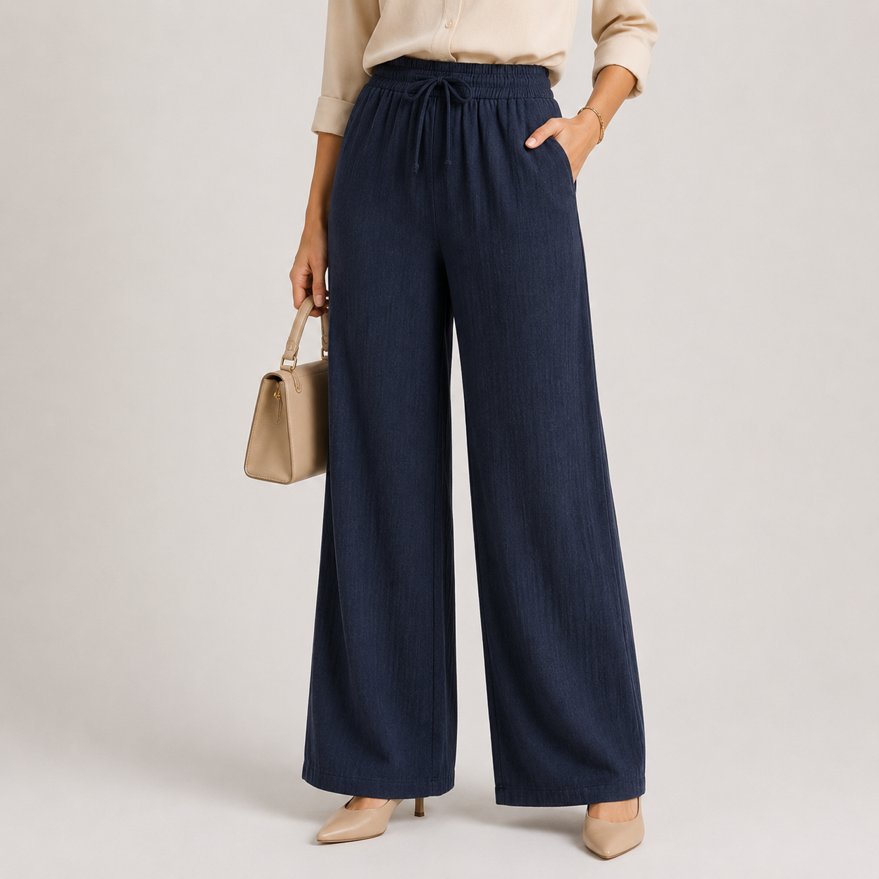 A model wearing dark navy blue wide-leg pleated trousers with a drawstring waist, paired with a beige blouse, beige heels, and holding a beige handbag against a light gray studio background.