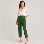 A woman modeling a pair of dark green cropped trousers with a cream button-down shirt and beige loafers, standing against a light gray studio background.