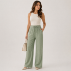 A woman modeling high-waisted, wide-leg sage green trousers with a drawstring waist, paired with a cream sleeveless top and beige heels, holding a small beige handbag against a light gray studio background.
