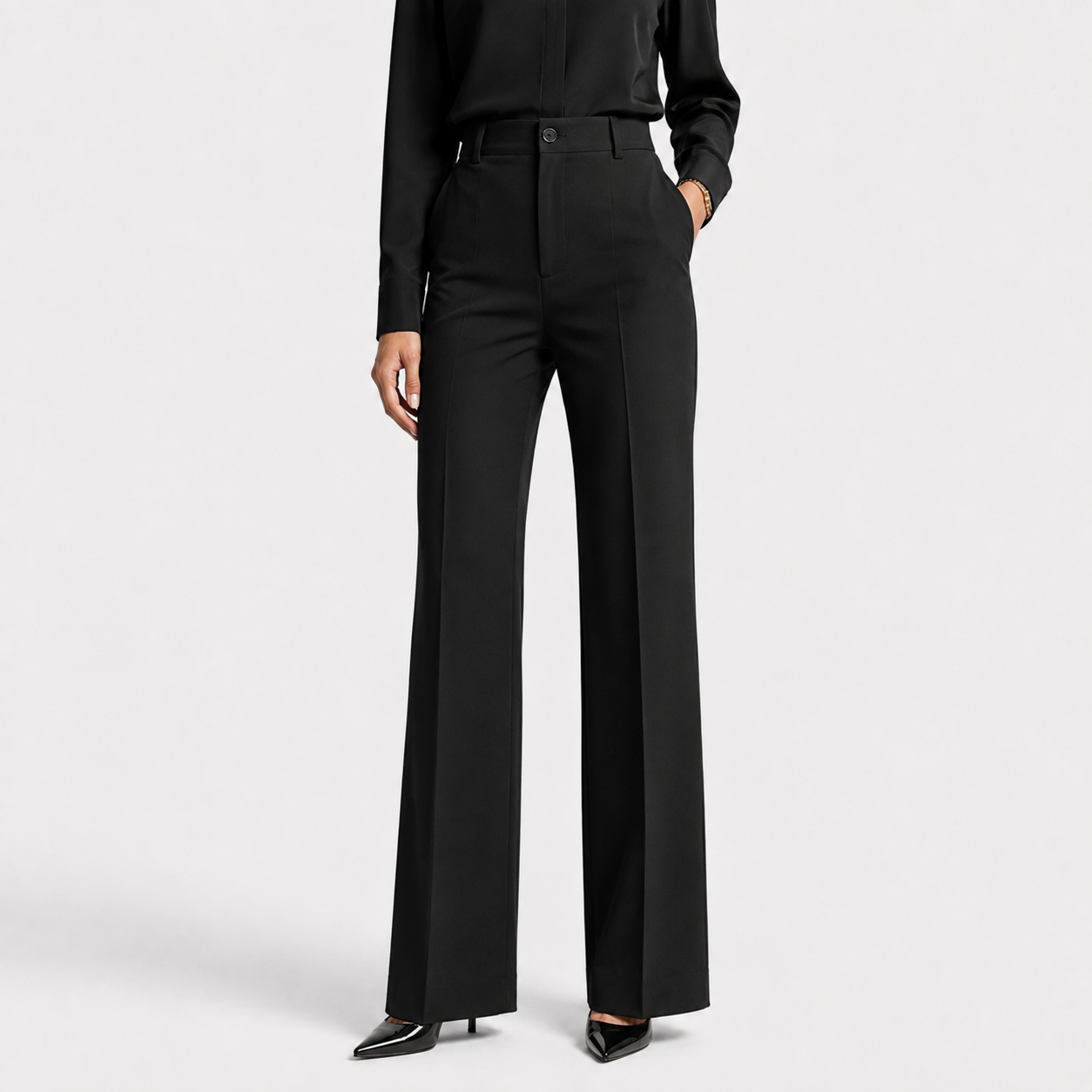 A model wearing black wide-leg trousers with a high waist, paired with a black long-sleeve top and black pointed-toe heels, shown against a light gray studio background.