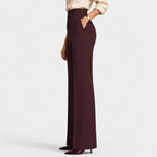 A model is shown from the waist down wearing high-waisted, wide-leg trousers in a deep burgundy color, paired with a light beige blouse and dark burgundy heels, against a plain light gray studio background.