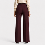 A model shown from the back wearing high-waisted, wide-leg trousers in a deep burgundy color, paired with a light beige blouse and black heels, against a plain light gray studio background.