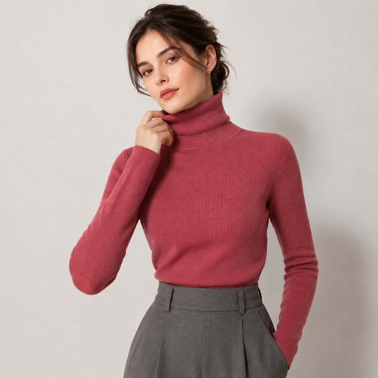 A woman wearing a ribbed turtleneck sweater in dusty rose pink, paired with gray trousers, posing against a neutral light gray background.