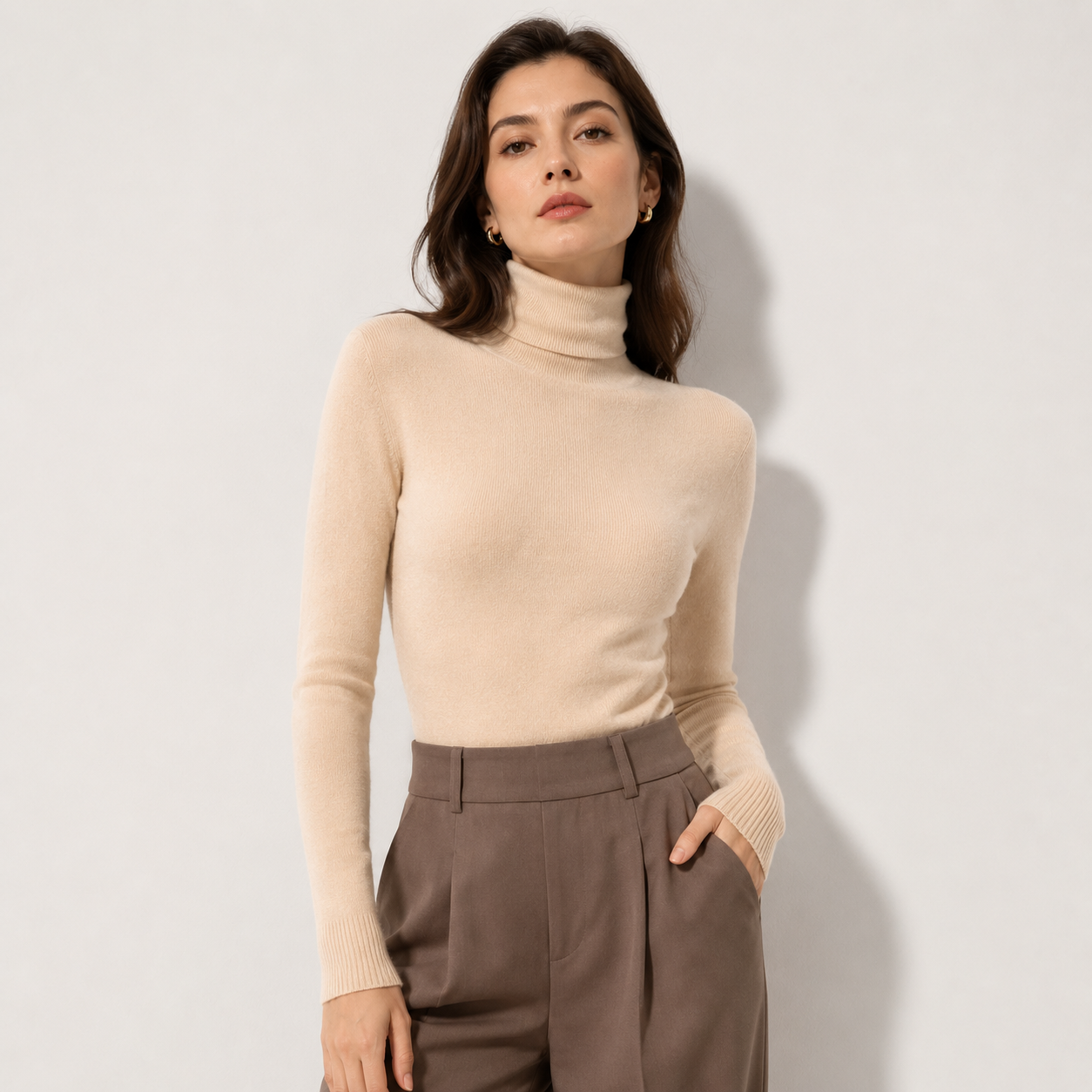 A woman modeling a beige ribbed turtleneck sweater paired with brown trousers, standing against a light gray studio background.