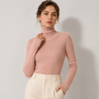 A woman wearing a ribbed pink turtleneck sweater and cream-colored trousers, standing against a light gray studio background.