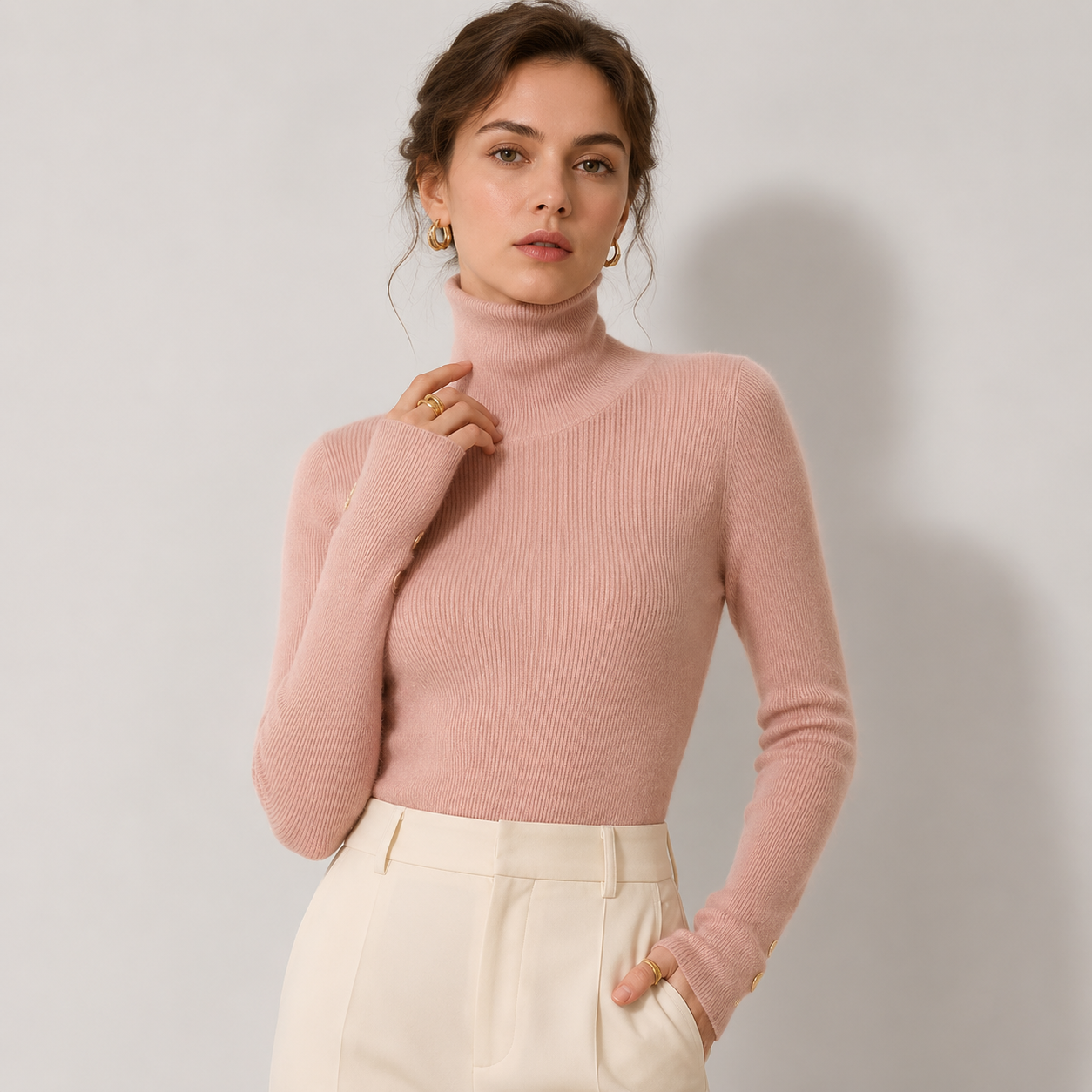 A woman wearing a ribbed pink turtleneck sweater and cream-colored trousers, standing against a light gray studio background.