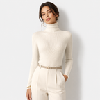 A woman modeling a fitted, ribbed cream turtleneck sweater paired with matching cream trousers and a beige belt, set against a plain light gray studio background.