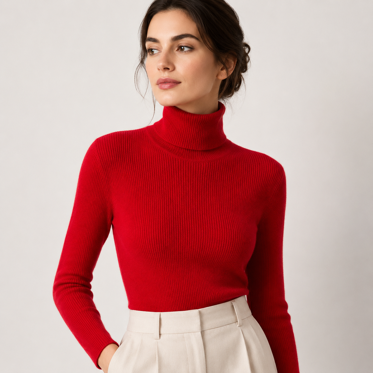 A woman wearing a bright red ribbed turtleneck sweater and cream-colored trousers, photographed against a neutral light gray studio background.