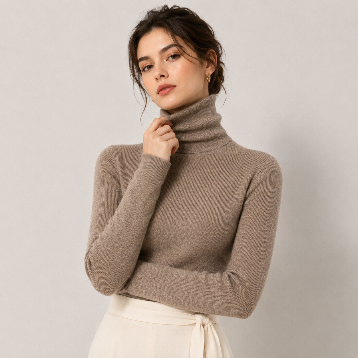 A woman wearing a taupe brown ribbed turtleneck sweater and a cream tie-waist skirt, posing against a neutral light gray background.