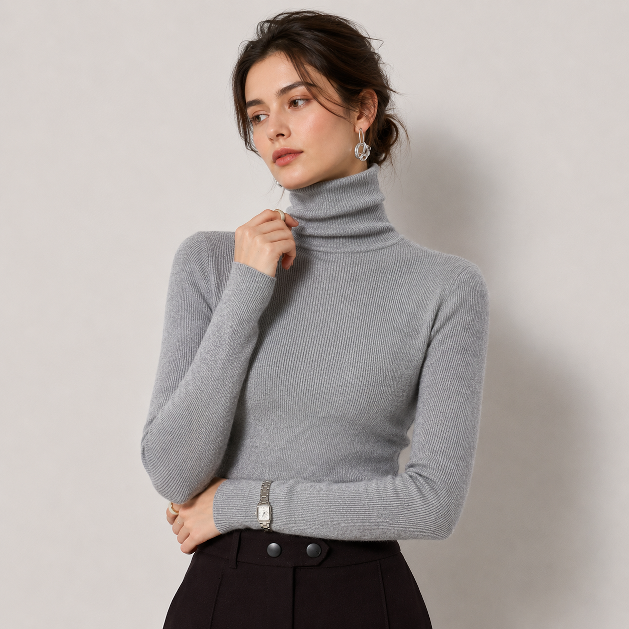 A woman wearing a light gray ribbed turtleneck sweater and dark trousers, posing against a neutral light gray background.