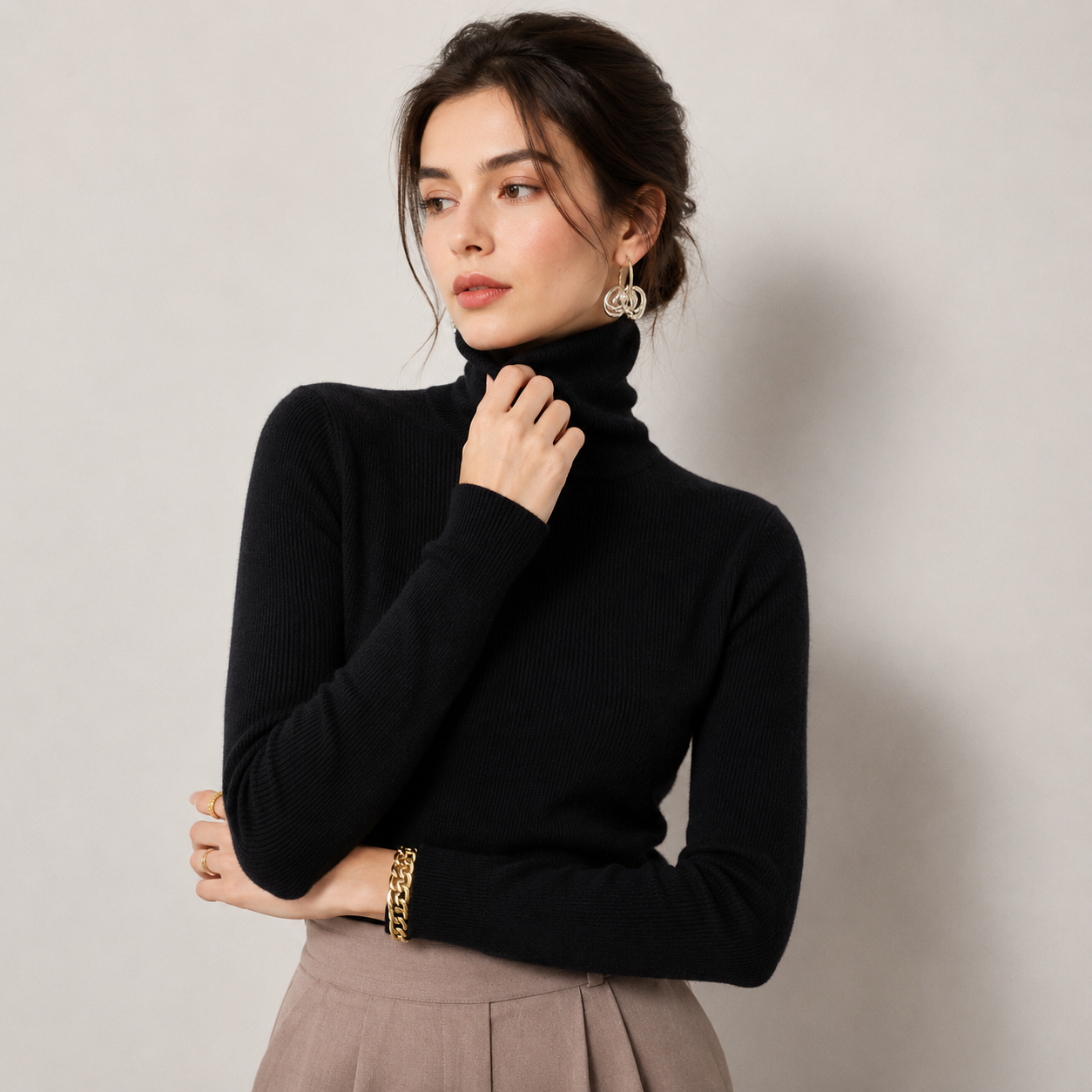 A woman wearing a black ribbed turtleneck sweater and a taupe pleated skirt, posing against a neutral light gray background.
