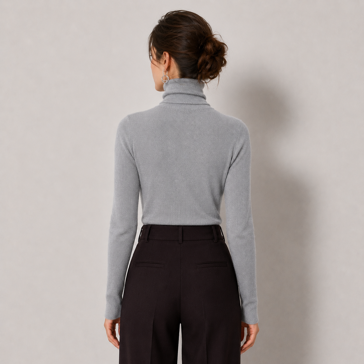 A woman shown from the back wearing a light gray ribbed turtleneck sweater and dark trousers, standing against a plain light gray studio background.