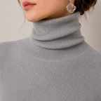 Close-up of a person wearing a light gray ribbed turtleneck sweater, with part of the face, neck, and hair visible. A clear geometric dangle earring is also visible against a plain light background.