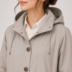 A close-up of a woman wearing a light taupe hooded jacket over a white collared shirt, cropped from the lower face to the upper torso against a plain light gray studio background.