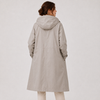 A woman shown from the back wearing a light beige hooded coat over cream pants, photographed against a plain light gray studio background.