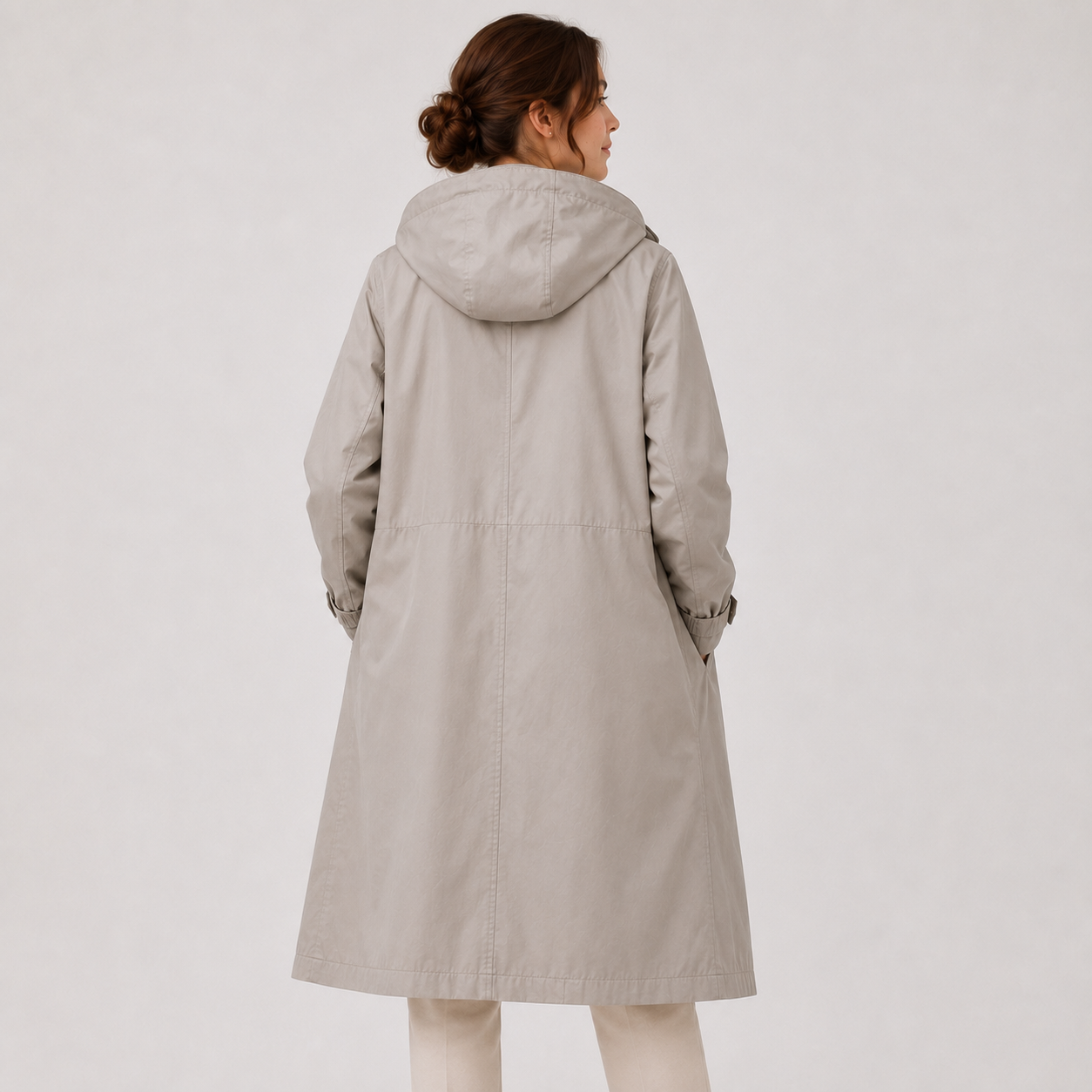 A woman shown from the back wearing a light beige hooded coat over cream pants, photographed against a plain light gray studio background.