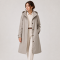 A woman modeling a mid-length light gray hooded trench coat over a cream shirt and cream trousers, standing against a plain light gray studio background.
