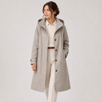 A woman modeling a mid-length light gray hooded trench coat over a cream shirt and cream trousers, standing against a plain light gray studio background.